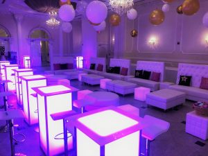 LED purple lit tables with metal chairs and white couches, black and gold pillows, underneath white, black, and gold balloons